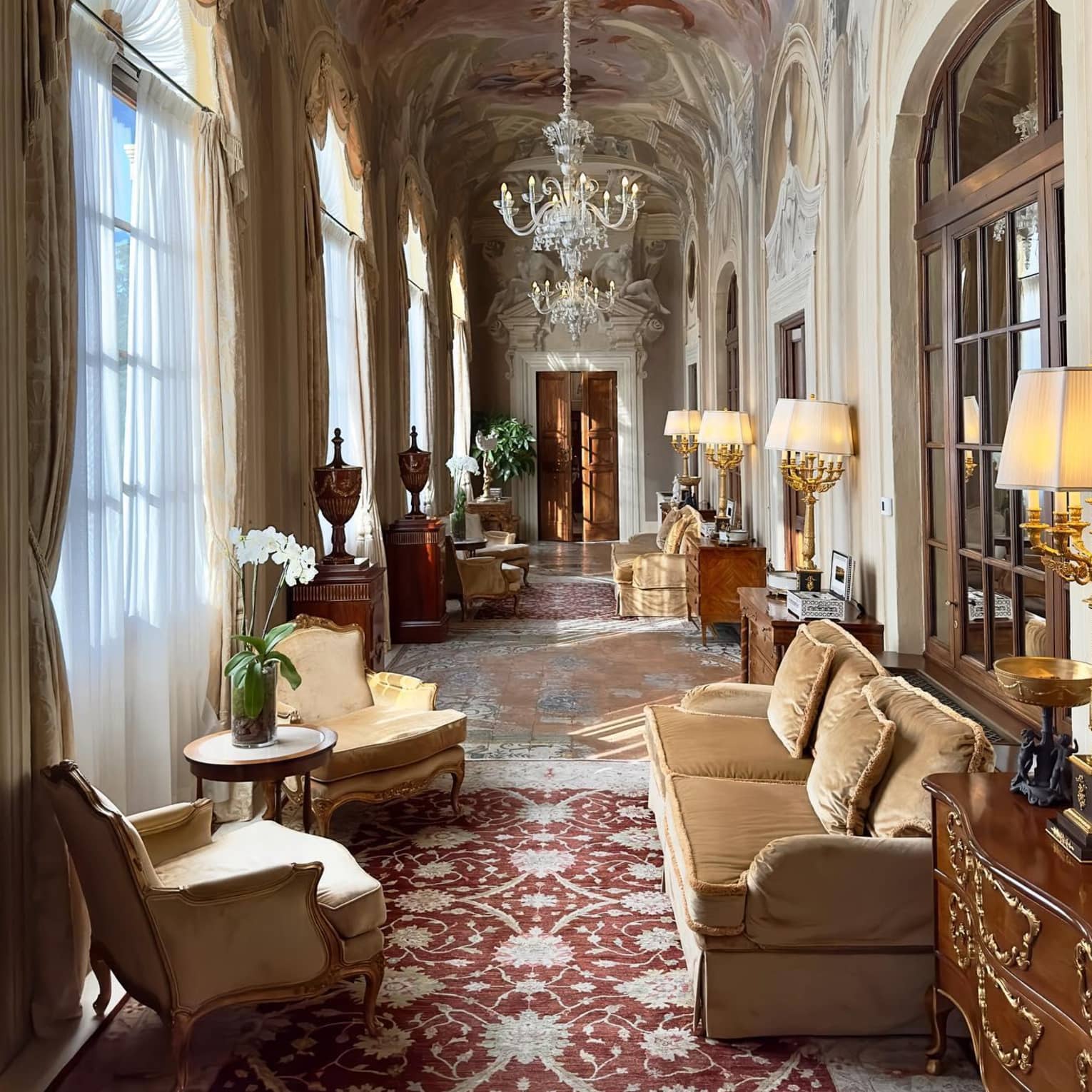 Luxurious hallway with ornate chandeliers, frescoed ceiling, large windows and plush seating, adorned with elegant lamps and decorative furniture.